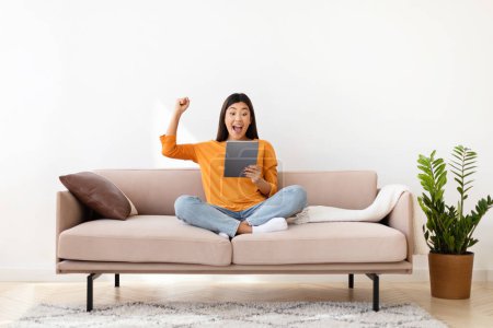 Photo for Smiling millennial asian lady holding digital tablet, gesturing, raising hand up, sitting on couch, home interior. Filipino woman reading great news, got cashback, copy space, white wall background - Royalty Free Image