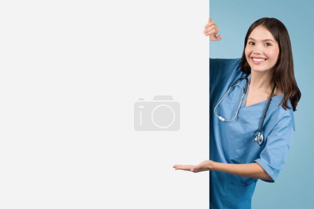 Foto de Enfermera joven profesional en abrigo azul apuntando hacia un lado en el espacio libre en la pizarra blanca en blanco, ideal para anuncios médicos, contra fondo azul vivo, pancarta - Imagen libre de derechos