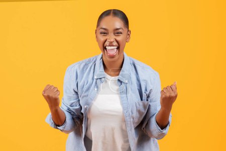 Photo for Yes, achievement. Joyful emotional black woman cheering and screaming shaking clenched fists, celebrating victory over yellow studio backdrop. I did it, great news concept - Royalty Free Image