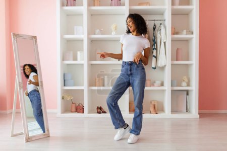 Photo for Joyful woman in white top and blue jeans dancing and enjoying her reflection in large mirror in room with pink walls and modern decor - Royalty Free Image