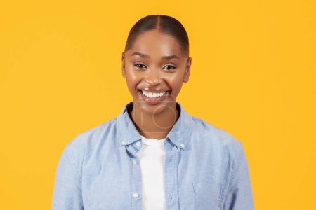 Téléchargez les photos : Portrait rapproché d'une femme noire heureuse en tenue décontractée en denim pose avec confiance, offrant un sourire joyeux, en studio sur fond jaune, incarnant un style millénaire confiant et la beauté - en image libre de droit