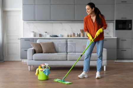 Photo for Delighted woman in vibrant orange shirt and yellow gloves mopping hardwood floor, with bucket filled with cleaning essentials beside her in a bright room - Royalty Free Image