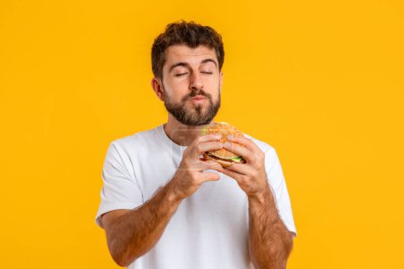 Photo for Junk Food. Hungry European Young Man Eating Tasty Burger, Standing Against Yellow Studio Backdrop. Guy Enjoying Unhealthy Comfort Food. Unbalanced Nutrition And Cheat Meal - Royalty Free Image