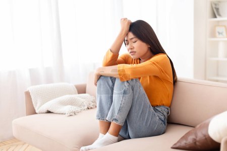 Photo for Upset young asian woman sitting on couch with legs up, touching her head, suffering from migraine during pms or period. Sad chinese lady feeling lonely, depressed, frustrated - Royalty Free Image