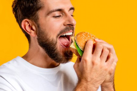 Photo for Closeup Portrait Of Joyful Millennial Man Biting Big Burger On Yellow Studio Backdrop, Having Food Cravings And Cheat Meal During Diet. Guy Eats Tasty Cheeseburger. Concept Of Junk Food - Royalty Free Image