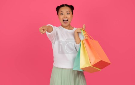 Foto de Joven dama asiática alegre con bolsas de compras apuntando a la cámara, mujer coreana alegre con compras seleccionándote, de pie sobre fondo de estudio rosa, disfrutando de las ventas de temporada, espacio de copia - Imagen libre de derechos