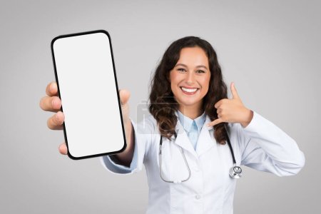Foto de Mujer feliz médico en abrigo que muestra el teléfono inteligente con pantalla en blanco, llámame signo con la mano, aislado en fondo gris estudio. Comunicación, aplicación, recomendación del sitio web para la atención médica - Imagen libre de derechos