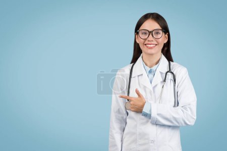 Photo for Optimistic young female doctor wearing glasses and white lab coat with stethoscope, pointing to her right at copy space with friendly smile, against blue backdrop - Royalty Free Image