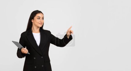 Téléchargez les photos : Jeune femme d'affaires caucasienne en costume avec tablette, pointant du doigt l'espace de copie, utiliser un écran virtuel, isolé sur fond gris. Planification du travail d'entreprise, démarrage avec l'appareil - en image libre de droit