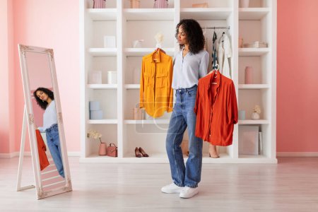 Téléchargez les photos : Femme latine réfléchie en jeans contemplant des choix de mode, tenant une chemise jaune et rouge, avec son reflet dans un miroir dans une pièce rangée - en image libre de droit