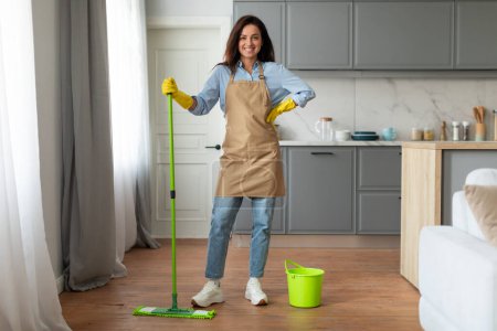 Photo for Cheerful young woman in beige apron, blue shirt, and jeans, holding mop beside green bucket, posing happily during a home cleaning session, full length - Royalty Free Image