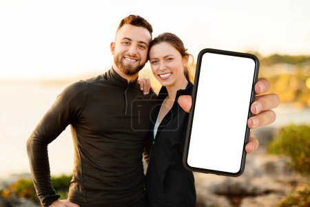 Téléchargez les photos : Convient et heureux couple tenant smartphone avec écran blanc, idéal pour la promotion de l'application, sur fond côtier flou pendant le coucher du soleil, maquette - en image libre de droit
