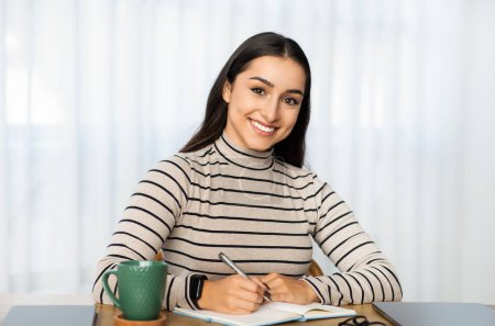 Foto de Mujer confiada del Medio Oriente joven con una sonrisa encantadora sentada en casa, escribiendo en su cuaderno con una taza verde y una tableta sobre la mesa, exudando productividad y satisfacción - Imagen libre de derechos