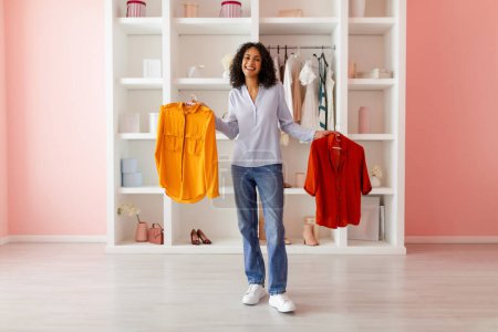 Photo for Happy woman in light room, holding yellow blouse and red one, contemplating her outfit choices in front of clean, organized wardrobe - Royalty Free Image