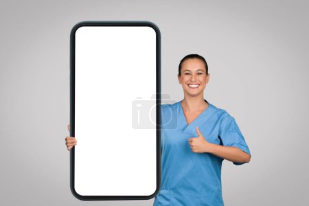 Téléchargez les photos : Femme heureuse médecin en manteau bleu tenir un téléphone énorme avec écran vide et montrant pouce geste vers le haut sur fond gris. Application médicale, recommandation du site Web - en image libre de droit