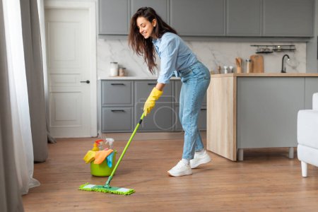 Téléchargez les photos : Femme engagée en tenue décontractée avec des gants jaunes nettoyant le sol en bois dans une cuisine bien éclairée, seau vert avec des fournitures de nettoyage à côté d'elle - en image libre de droit