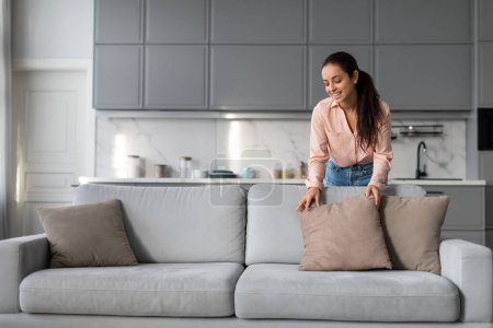 Photo for Content woman meticulously arranges decorative pillows on stylish couch, adding the finishing touches to her well-maintained contemporary living space - Royalty Free Image