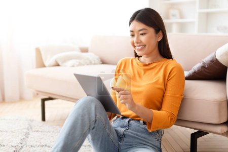 Photo for Happy long-haired young asian woman sitting on floor, reading news on digital tablet, watching video content online, copy space. Chinese lady enjoying weekend at home, scrolling on pad - Royalty Free Image