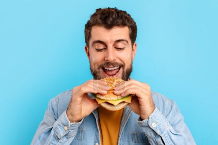 Photo for Junk Food Overeating. Happy caucasian man eats burger and smiling, enjoying tasty unhealthy snack, standing over blue studio background, portrait shot. Cheat meal, nutrition and male diet concept - Royalty Free Image