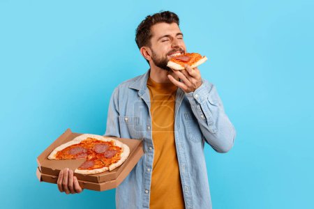 Téléchargez les photos : Jeune homme barbu mangeant une tranche de pizza debout posant avec la boîte délivrée de la pizzeria sur fond bleu studio. Mangeur de malbouffe Profitant d'un repas malsain Cheat. Nutrition masculine et suralimentation - en image libre de droit
