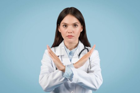 Photo for Young female doctor in white coat sternly displays stop gesture with her hands, looking at camera against plain blue background, signifying caution - Royalty Free Image