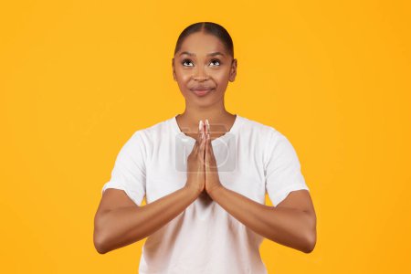 Foto de Fe. Retrato de la sonriente dama afroamericana orando con las manos apretadas, la joven religiosa negra de pie y orando con esperanza sobre el fondo del estudio amarillo - Imagen libre de derechos