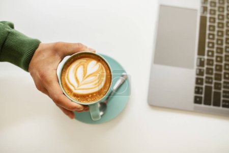 Téléchargez les photos : Vue du dessus du mec noir buvant du café et travaillant sur ordinateur au café. Coupé de la main de l'homme prenant tasse avec cappuccino tout en utilisant un ordinateur portable, assis à la table blanche - en image libre de droit