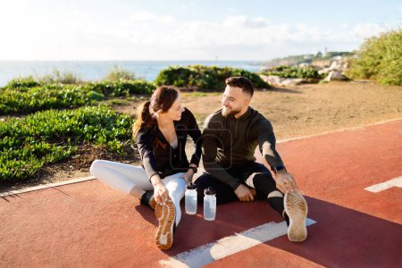 Foto de Hombre y mujer gozosos en ropa deportiva sentados y estirados en pista de atletismo con botellas de agua, compartiendo un momento de bienestar y risa - Imagen libre de derechos