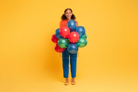 Téléchargez les photos : Joyeuse adolescente noire sourit alors qu'elle tient un tas de ballons lumineux et festifs debout sur fond de studio jaune. Fête de vacances et concept de célébration d'anniversaire. Plan complet - en image libre de droit