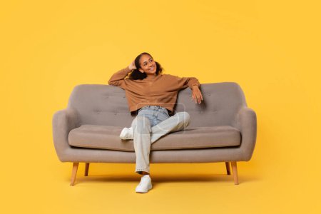 Foto de Relajada adolescente negra estudiante descansando en el sofá cómodamente, disfrutando del descanso expresando felicidad y relajación casual sobre el fondo del estudio amarillo. Disparo completo - Imagen libre de derechos