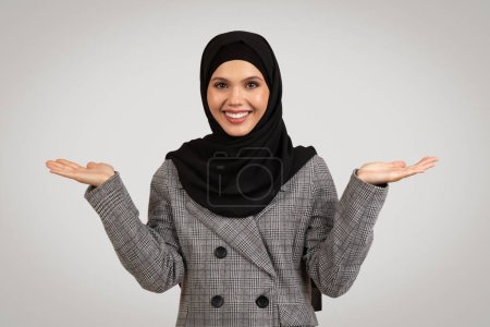 Foto de Mujer profesional sonriente en elegante chaqueta houndstooth y hijab negro hace gesto de equilibrio con las manos abiertas contra fondo gris neutro - Imagen libre de derechos