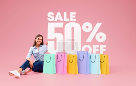 Téléchargez les photos : Offre Shopping Discount. Souriant European Shopaholic Woman Publicité 50 pour cent vente assis sur le sol avec des sacs à provisions colorés, sur fond de studio rose. Collage - en image libre de droit