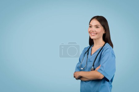 Photo for Cheerful young nurse in blue coat standing confidently with arms folded and looking at free space, her smile suggesting friendliness and competence, against blue backdrop - Royalty Free Image
