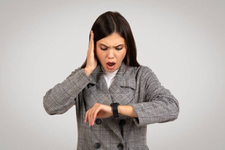 Foto de Preocupada joven profesional mirando su reloj inteligente con la mano en la cabeza, indicando estrés o llegar tarde, vistiendo traje de perro, fondo gris - Imagen libre de derechos
