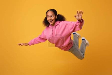 Photo for Happy carefree African American teenager girl in casual flying joyfully with hands spread, exuding freedom and fun on yellow studio backdrop, posing in mid air. Full length shot - Royalty Free Image