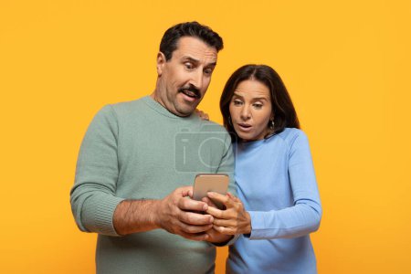 Photo for Surprised old european couple looking at smartphone screen, man holding the device in hands, woman reacting with shock, both standing against a vibrant yellow studio backdrop - Royalty Free Image