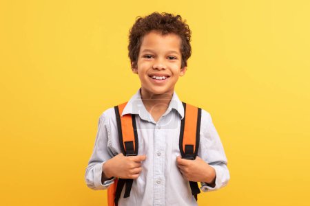 Foto de Amable colegial latino con cabello rizado y sonrisa brillante, con camisa blanca y mochila naranja, de pie contra alegre fondo amarillo - Imagen libre de derechos