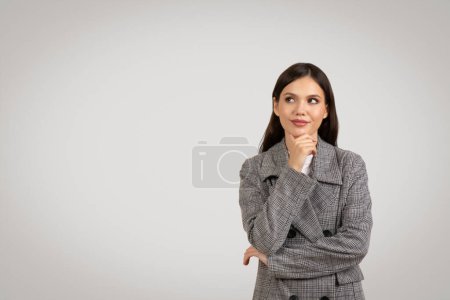 Téléchargez les photos : Jeune femme pensive en veste à carreaux gris repose son menton sur la main, regardant vers le côté à l'espace libre, perdu dans la pensée sur fond gris doux - en image libre de droit