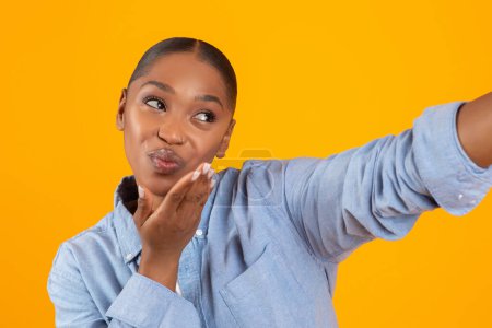 Téléchargez les photos : Flirty afro-américaine Lady souffler un baiser faisant Selfie, Flirtant dans les médias sociaux ou Sortir ensemble application, Posant sur fond jaune dans Studio. Portrait De Jolie Femme Noire Envoi D'air Embrasser - en image libre de droit