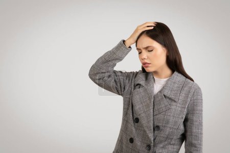 Photo for Worried young woman in blazer holds her head, expressing confusion or headache, with troubled look against neutral grey background - Royalty Free Image
