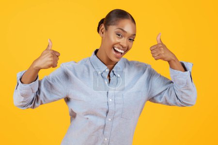 Téléchargez les photos : Le pouce levé. Dame afro-américaine excitée montrant comme geste signe souriant à la caméra sur fond jaune studio. Femme noire en denim occasionnel chemise approuvant l'offre de publicité - en image libre de droit
