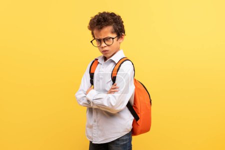 Foto de Escolar serio con gafas y mochila naranja, de pie con los brazos cruzados y mirando a la cámara sobre fondo amarillo sólido - Imagen libre de derechos