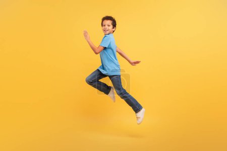 Photo for Energetic black boy captured in mid-run with joyful expression, sporting blue t-shirt and jeans, set against vivid yellow background - Royalty Free Image