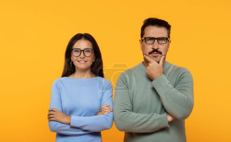 Photo for A contemplative caucasian man and a confident woman stand side by side, arms crossed, with the man touching his chin in thought and the woman smiling against an orange backdrop - Royalty Free Image