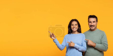 Téléchargez les photos : Un homme et une femme caucasiens heureux portant des vêtements décontractés pointent sur le côté avec des sourires lumineux, indiquant un objet ou un concept, sur un fond orange monochrome - en image libre de droit