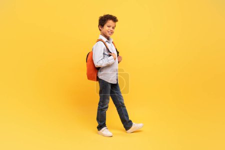 Photo for Smiling schoolboy with curly hair, carrying orange backpack and walking confidently against bright yellow background, suggesting movement and energy - Royalty Free Image