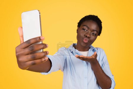 Foto de Joven bloguera estudiante negra positiva enviando beso aéreo, hacer selfie en el teléfono, aislado en el fondo amarillo, estudio. Blog, redes sociales, videollamadas y gestos de amor - Imagen libre de derechos