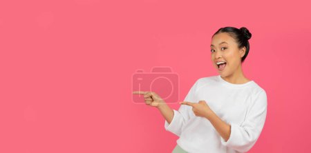 Photo for Joyful asian woman pointing aside at copy space, happy korean female standing isolated over pink studio background, signaling new opportunity, advertising new offer or great promo, panorama - Royalty Free Image