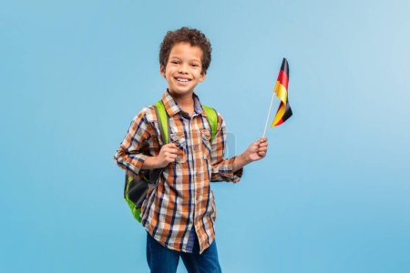 Téléchargez les photos : Garçon intelligent en chemise à carreaux, tenant joyeusement le drapeau allemand, avec un sac à dos vert sur les épaules, sur un fond bleu vif - en image libre de droit