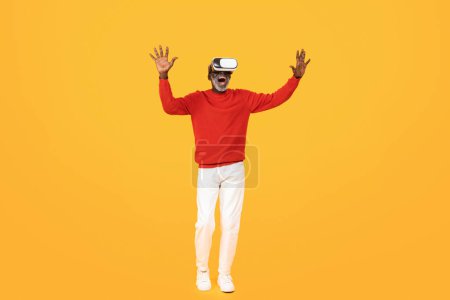Photo for Cheerful shocked old african american man in vr 3d glasses, with open mouth, control virtual screen with hands, isolated on orange background, full length. Entertainment, fun and game - Royalty Free Image